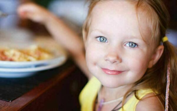 A little girl smiles and looks at the camera while raising a utensil in her hand as she sits at a table eating a meal.