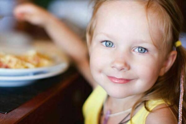 A little girl smiles and looks at the camera while raising a utensil in her hand as she sits at a table eating a meal.