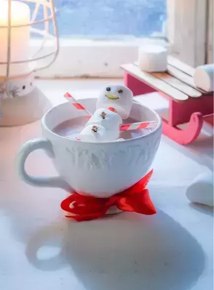 White mug of hot chocolate with a marshmallow snowman "soaking," propped by red & white candy cane "arms". The snowman has a smiley face. A red ribbon is tied around the mug. A lit lantern and a miniature wooden sled with marshmallows sit near a cold windowpane.