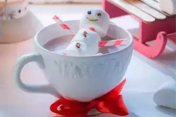 White mug of hot chocolate with a marshmallow snowman "soaking," propped by red & white candy cane "arms". The snowman has a smiley face. A red ribbon is tied around the mug. A lit lantern and a miniature wooden sled with marshmallows sit near a cold windowpane.