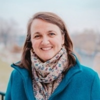 Smiling woman with brown hair, blue eyes, wearing a teal coat and a colorful patterned scarf, outdoors.