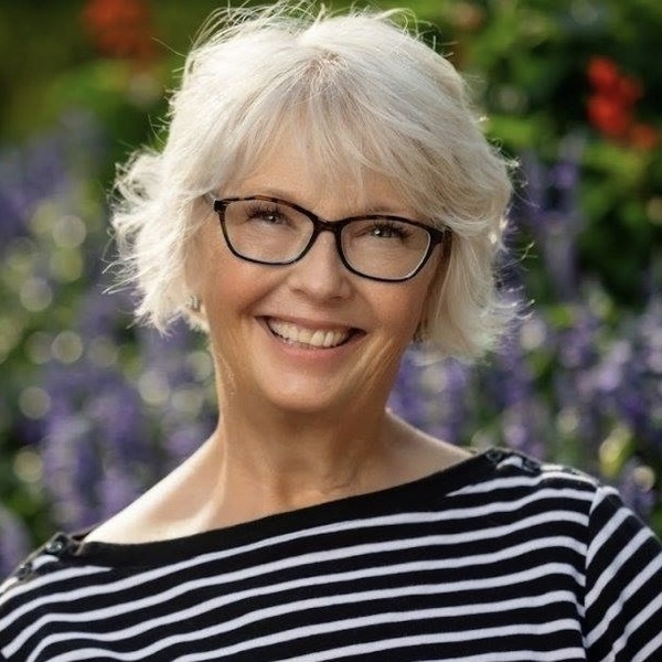 A smiling woman with short gray hair, dark-rimmed glasses, and a black and white striped shirt in a garden setting.