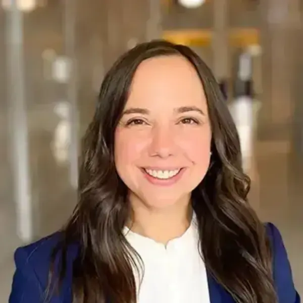 Jill Curry, a smiling woman with dark, wavy hair wears a white collared shirt and a navy blue blazer in a blurred indoor setting.